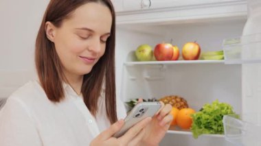 Gülümseyen kahverengi saçlı, beyaz tişörtlü çekici bir kadın buzdolabından portakal alan akıllı telefonunu kullanarak taze meyve ve sebzelerle dolu açık buzdolabının yanında duruyor.
