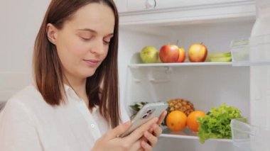 Beyaz tişörtlü beyaz bir kadın açık buzdolabının yanında duruyor. Süpermarketten satın alma listeleri yazan mobil kalori uygulamalarını kontrol ediyor.