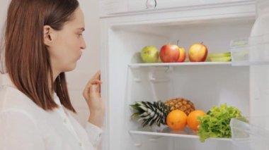 Kahverengi saçlı, hayal kırıklığına uğramış beyaz kadın açık buzdolabının yanında duruyor ve iğrenç yeşil marul yapraklarıyla lezzetli, sağlıksız atıştırmalıklar arıyor.