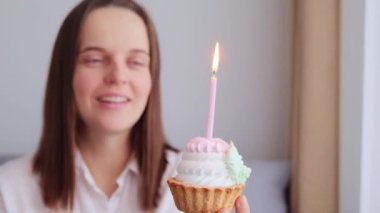 Aşırı neşeli siyah saçlı kadın elinde pastayla evde oturuyor. Gözlerinde dilek tutuyor. Doğum gününü kutlarken mutlu yüzüyle yanan mum üflüyor.