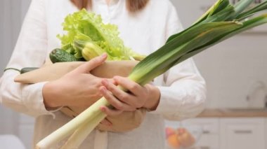 Beyaz gömlek giyen, elinde kağıt torba ve sebze tutan, salata hazırlamak için pırasa ve evde diğer sağlıklı yemekleri gösteren kimliği belirsiz bir kadın.