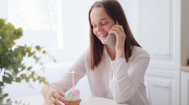 Gülümseyen, beyaz tişörtlü genç bir kadın. Doğum gününü kutluyor. Mutfakta pasta ve mumla oturuyor.