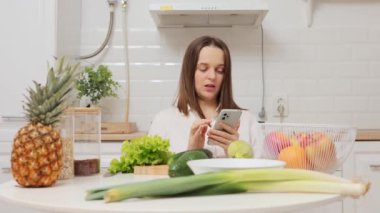 Sağlıklı beslenme tarifi için akıllı telefon aracılığıyla meyve ve sebzelerle dolu bir masada oturup web sayfalarını karıştıran konsantre beyaz kadın.