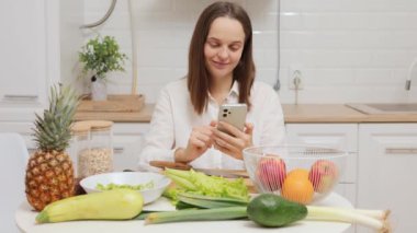 Gülümseyen mutlu beyaz kadın cep telefonunu kullanıyor. Mutfakta meyve ve sebzelerle dolu bir masada oturuyor. İnternette kayıyor.
