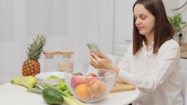 Kahverengi saçlı beyaz kadın akıllı telefon kullanıyor. Mutfakta meyve ve sebzelerle dolu bir masada oturuyor. Lezzetli organik yemek hazırlıyor.