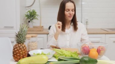 Koyu renk saçlı, beyaz tişörtlü beyaz kadın mutfakta meyve ve sebzelerle birlikte yeşil salata yiyerek oturuyor.
