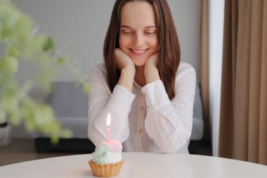 Neşeli, pozitif, neşeli, beyaz, kahverengi saçlı kadın doğum gününü pasta ve mumla kutluyor. Tatlısına bakıyor, mutlu bir gülümsemeyle, dileğini düşünüyor.