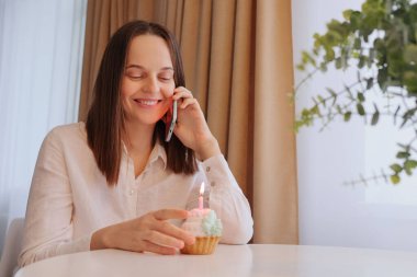 Beyaz gömlekli neşeli bir kadın evde oturmuş elinde mumla telefonda konuşuyor. Doğum gününü kutluyor. Mobil tebriklerin tadını çıkarıyor.