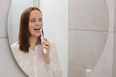 Dişlerine dikkat et. Banyo dişçisinin temel ihtiyaçları. Ağız sağlığı rutini. Taze ve temiz bir nefes. Gelişmiş oral sağlık. Beyaz, genç, yetişkin bir kadın aynanın önünde dişlerini fırçalıyor.