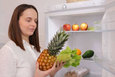 Meyve ve sebzelerle dolu açık buzdolabının yanında duran tatlı beyaz kadın taze ananas ve yeşil marul yapraklarıyla ne pişireceğini düşünüyor.