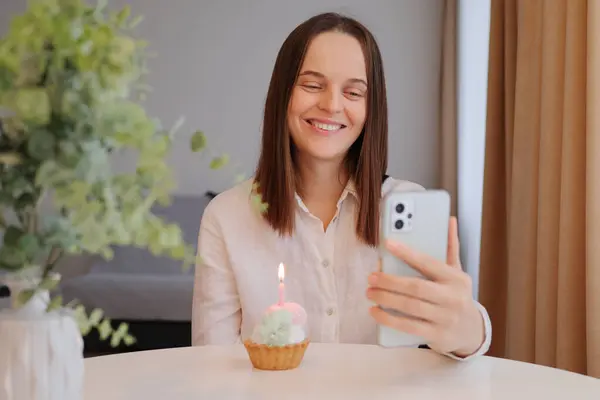 Smiling cheerful Caucasian woman with birthday cake and smartphone in home interior celebrating her holiday with her friend online via video call looking at mobile phone screen