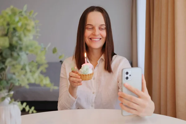 Joyful Caucasian woman with birthday cake and phone in home interior showing her dessert to cell phone screen talking via video call with friend enjoying online celebration
