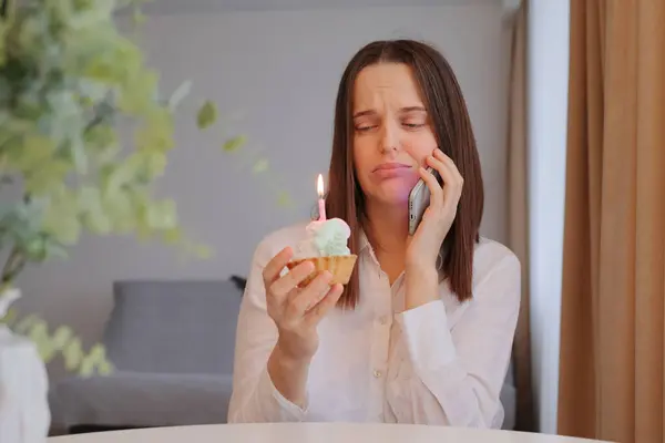 Unhappy sad upset woman with dark hair sitting at table at home holding cake with candle and talking with mobile phone expressing negative emotions feeling loneliness