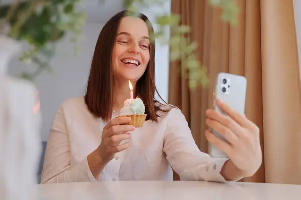 Smiling woman with cake and burning candle celebrating birthday on video call holding smartphone in hands enjoying festive event being in good mood listening congratulations
