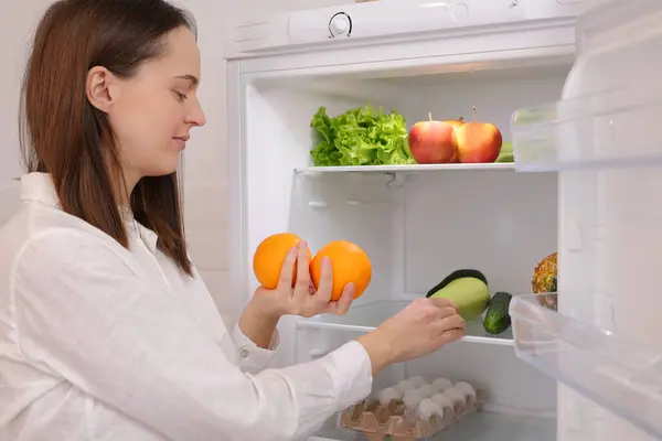 Caucasian woman standing near open refrigerator full of fruits and vegetables choosing fruits prefers organic nutrition female preparing to cook enjoying healthy eating