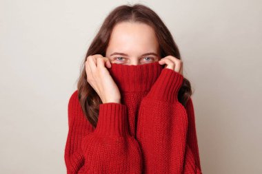 Kahverengi saçlı kadın kırmızı moda süveterini kafasına geçirip eğleniyor. Çocuksu kıyafetleriyle ortadan kayboluyor. Gri arka planda gizlenmiş neşeli utangaç gözlerle altından bakıyor.