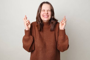 Kahverengi saçlı, kahverengi kazaklı çekici bir kadın. Gri arka planda yalnız poz veriyor. Parmakları çapraz ve gözleri kapalı iyi şanslar dilemek için dua ediyor.