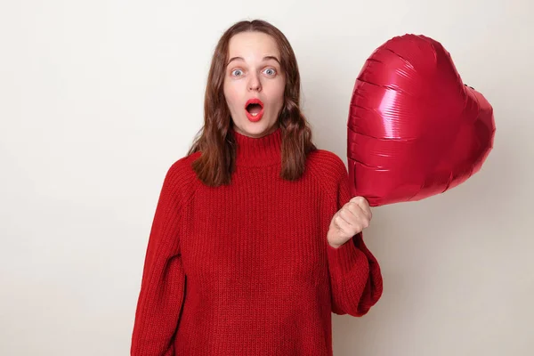 Shocked astonished Caucasian woman in red sweater holding heart shaped balloon isolated over gray background looking at camera with big eyes saying wow omg sees crush on blind date on Valentines Day