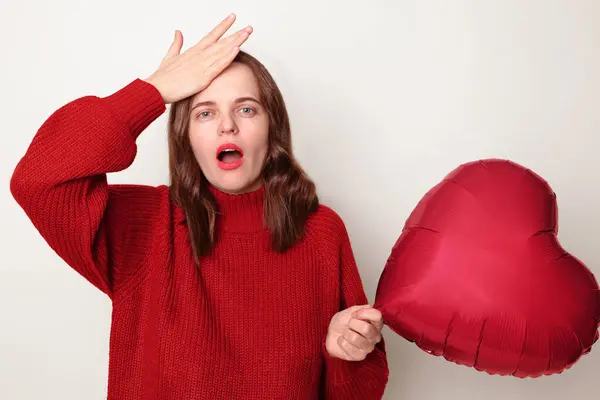 Shocked amazed woman with brown hair wearing red jumper standing isolated over gray background with heart shaped balloon showing facepalm gesture feeling sorrow regret blaming herself for failure