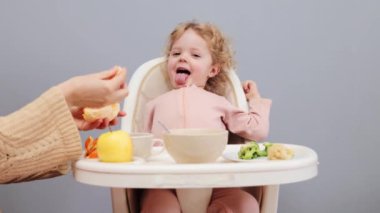 Küçük, sarışın, dalgalı saçlı kız. Gri arka planda tek başına tek başına yemek yiyor. Tanınmayan bir kadının eli, lezzetli meyvelerin tadını çıkaran çocuğa portakal veriyor.