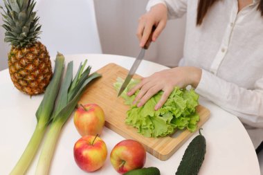 Mutfak masasında meyve ve sebzelerle oturan tanınmamış bir kadın sağlıklı salata için yeşil marul yaprakları kesiyor.