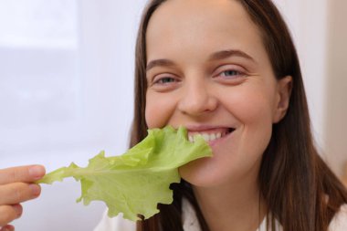 Mutfak arka planında yeşil salata yaprağını ısıran kahverengi saçlı, mutlu, beyaz kadın sağlıklı beslenmenin keyfini çıkarıyor.