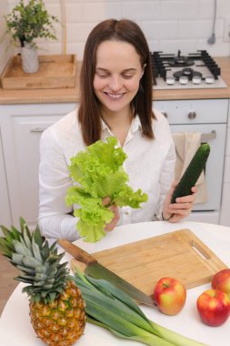 Mutfak masasında elinde meyve, sebze, salatalık ve yeşil marul yapraklarıyla oturan neşeli, beyaz saçlı kadın. Salata için organik malzemeler.