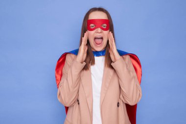 Heyecanlı kahverengi saçlı kadın süper kahraman kostümü giyiyor. Mavi arka planda yalnız poz veriyor. Elleri ağıza yakın duruyor.
