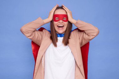 Komik, pozitif, neşeli, kahverengi saçlı, süper kahraman kostümü giyen, mavi arka planda yalnız poz veren, elleri başının üstünde duran ve gülen bir kadın.
