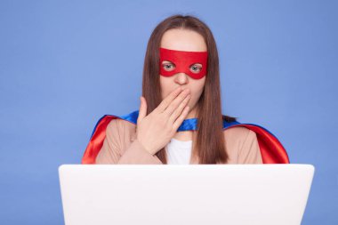 Şaşırdım. Kahverengi saçlı, süper kahraman kostümü giyen ve maske takan bir kadın mavi arka planda saklanıyor. Bilgisayarda çalışıyor, uzaktan kumandayla sorun yaşıyor.