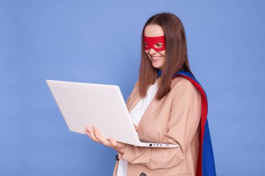 Kahverengi saçlı, süper kahraman kostümü giymiş ve maske takmış bir kadın mavi arka planda izole bir şekilde duruyor bilgisayardaki uzaktan kumandalı işiyle klavyede daktilo ekranına bakıyor.