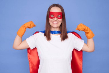 Neşeli, kahverengi saçlı, süper kahraman maskesi takan ve lastik eldiven takan kadın kollarını kaldırdı ve mavi arka planda izole bir şekilde poz verdi.