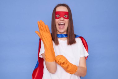 Aşırı mutlu, kahverengi saçlı, süper kahraman kostümlü ve maskeli bir kadın. Mavi arka planda izole bir şekilde poz veriyor. Turuncu lastik koruyucu eldivenler giyip etrafı toparlamaya hazırlanıyor.