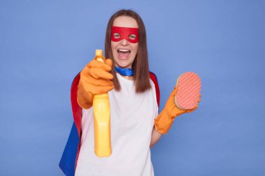 Heyecanlı kahverengi saçlı, süper kahraman maskesi takan ve koruyucu eldiven takan mavi arka planda izole edilmiş bir şekilde duran elinde deterjan spreyi ve süngerle temizliğe yardım etmeye hazır bir kadın.