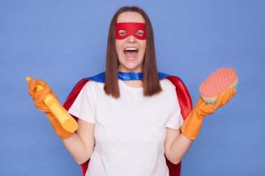 Süper kahraman maskesi takan çılgın bir kadın ve koruyucu lastik eldivenler giyip elinde deterjan ve süngerle mavi arka planda izole edilmiş poz vererek temizlik işindeki işinden zevk aldığını iddia ediyor.