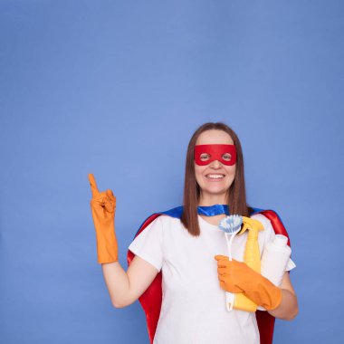 Gülümseyen, mutlu yetişkin kadın, eldiven giyiyor ve süper kahraman kostümü giyiyor. Elinde mavi arka planda izole edilmiş deterjan var. Reklam için fotokopi alanını işaret ediyor.