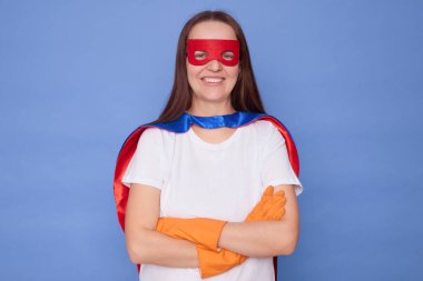 Kendine güvenen, beyaz saçlı, süper kahraman kostümü giyen beyaz tişört giyen, mavi arka planda izole edilmiş lastik eldiven takan, çapraz ellerle kameraya bakan, dişlek gülümsemeli bir kadın.