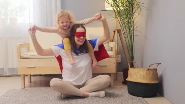 ママの肩に座っているスーパーヒーローの衣装の子供を着て一緒に遊んでいる彼女の小さな幼い娘と陽気な幸せな女性 — ストック動画