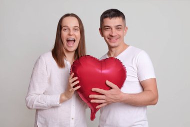 Gri arka planda izole edilmiş kalp şeklinde balonları olan heyecanlı çift kameraya neşe ve heyecanla bakıyor Sevgililer Günü 'nü kutluyorlar.