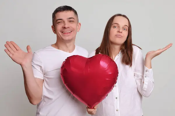 Confused puzzled man and woman wearing white clothing holding heart shaped air balloon isolated over gray background spreading hands do not know how to celebrate anniversary