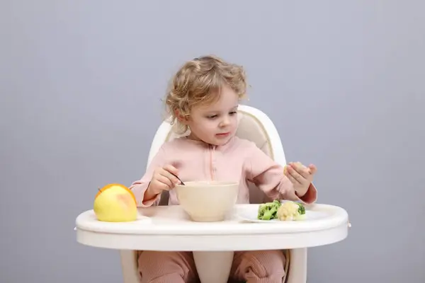 Little infant baby girl eating healthy food while sitting in high chair isolated over gray background taking broccoli having breakfast or dinner enjoying delicious dish