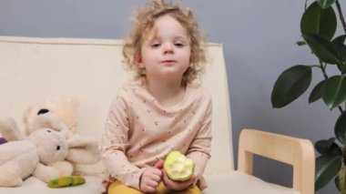 Çekici, küçük, sarışın, dalgalı, kanepede oturan ve evin içinde yeşil elma yiyen tatlı, lezzetli meyveli atıştırmalıkların tadını çıkaran bir kız.