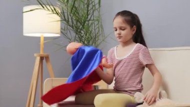 Koyu kahverengi saçlı, at kuyruklu güzel bir kız evinde oyuncak bebeğiyle oynarken süper kahraman kostümü giydiği ilginç maceralar hayal ediyor.