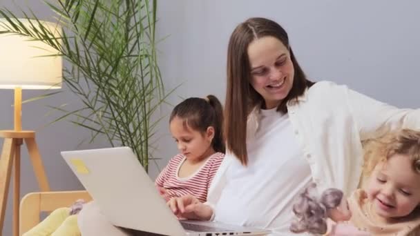 当孩子们的女儿们坐在休产假的妈妈旁边的沙发上玩洋娃娃时 年轻的笑容让妈妈们满意 她们用笔记本电脑做自由职业 — 图库视频影像