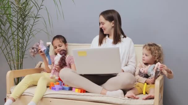 ハッピー陽気な白人ブラウンハイレッドの母親はラップトップで働いていますが 子供たちはキーボードにタイピングしながら家のインテリアの女性のおもちゃで遊んでいます — ストック動画
