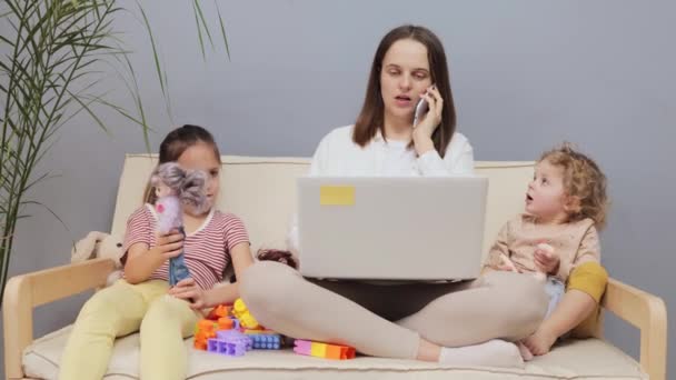 白种人的浓密棕色头发的母亲在笔记本电脑上工作 在手机上聊天 而她的孩子们在家里玩洋娃娃时 她正在做她休产假期间的远程工作 — 图库视频影像