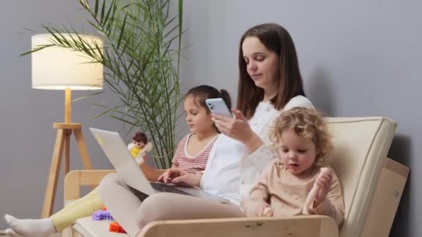 漂亮的白种人棕色头发的母亲一边在笔记本电脑上工作 一边用手机在网上打滚 而她的孩子们则在家里客厅里玩玩具 — 图库视频影像