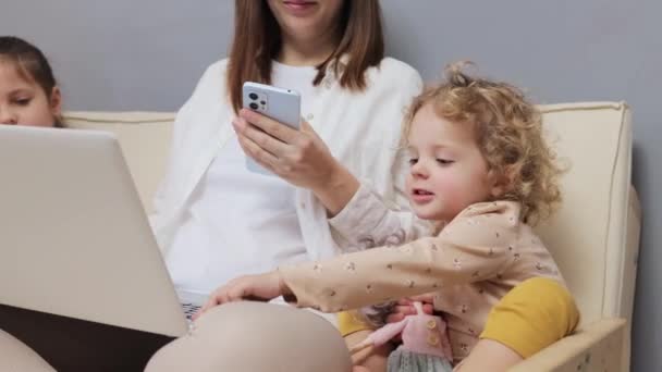 カカシアの茶色の毛髪の母親は スマートフォンでラップトップとブラウジングのウェブページで作業しながら 彼女の子供たちは リモートワークと出産休暇を組み合わせています — ストック動画