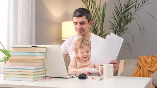 白种人忙碌的男人穿着白色的T恤 在笔记本电脑上工作 带着文件坐在桌旁 他的小女儿坐在家里 用遥控器和纸干扰着她的父亲 — 图库视频影像