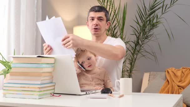 管理职业和家庭生活 虚拟时代的父母身份 与孩子的工作与生活的平衡 忙碌的男子穿着T恤衫 和他的幼儿一起在笔记本电脑上工作 他们的文件都放在家里的桌子上 — 图库视频影像